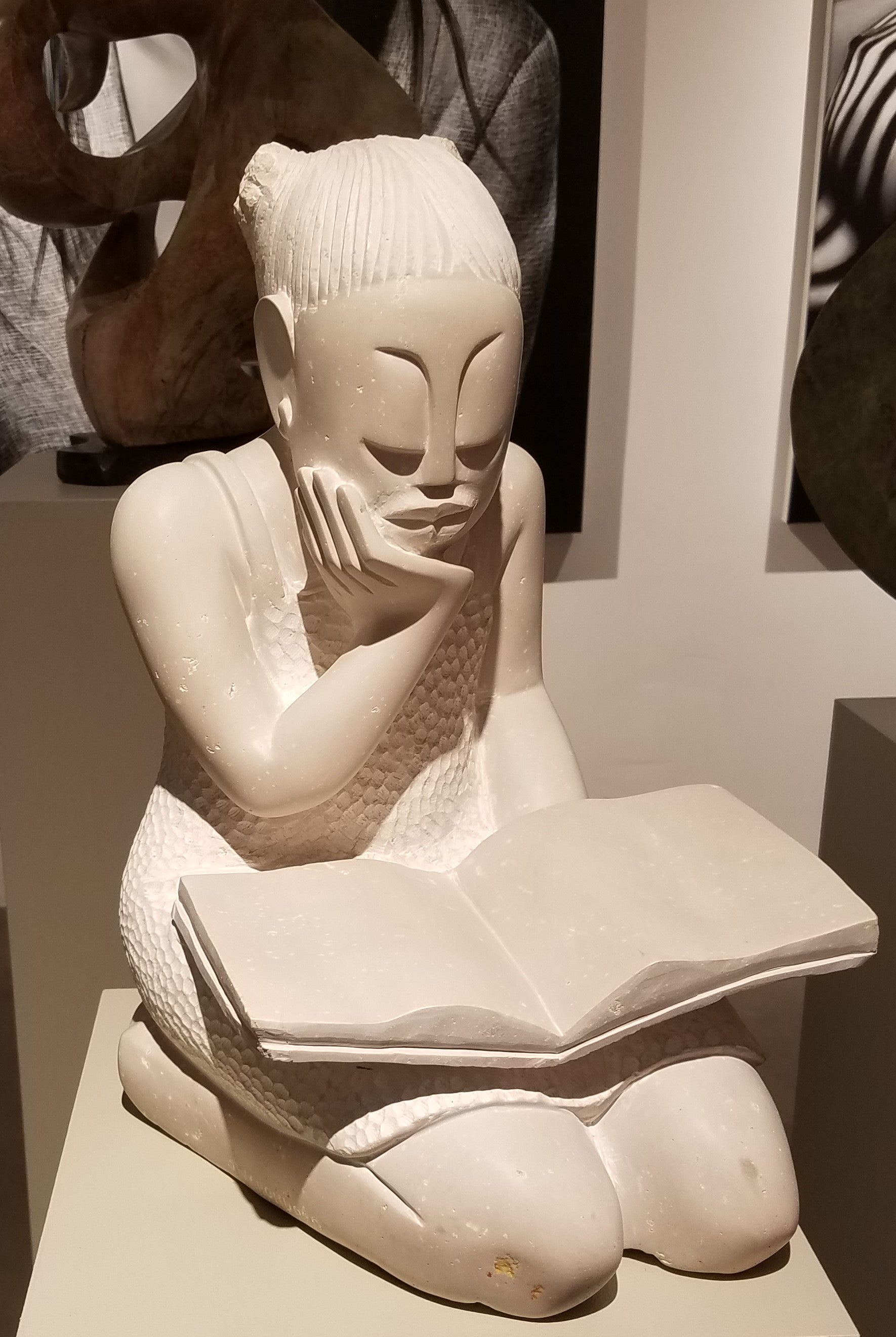 Stone sculpture of a girl reading a book. The stone color is white, with the more sanded and smooth areas being slightly a cream color, or off white. The dimensions are 16.5”H x 10”W x 11.5”D - 60 lbs.