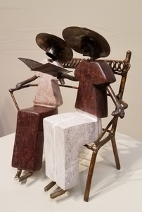Sculpture of a couple sitting on a bench reading together from the same book. It was created from a combination of rapoko stone (a soft serpentine), and metal, in Zimbabwe. The people’s bodies are sculpted from stone, and the bench, arms, legs, heads and hats, are all metal, welded and soldered together.