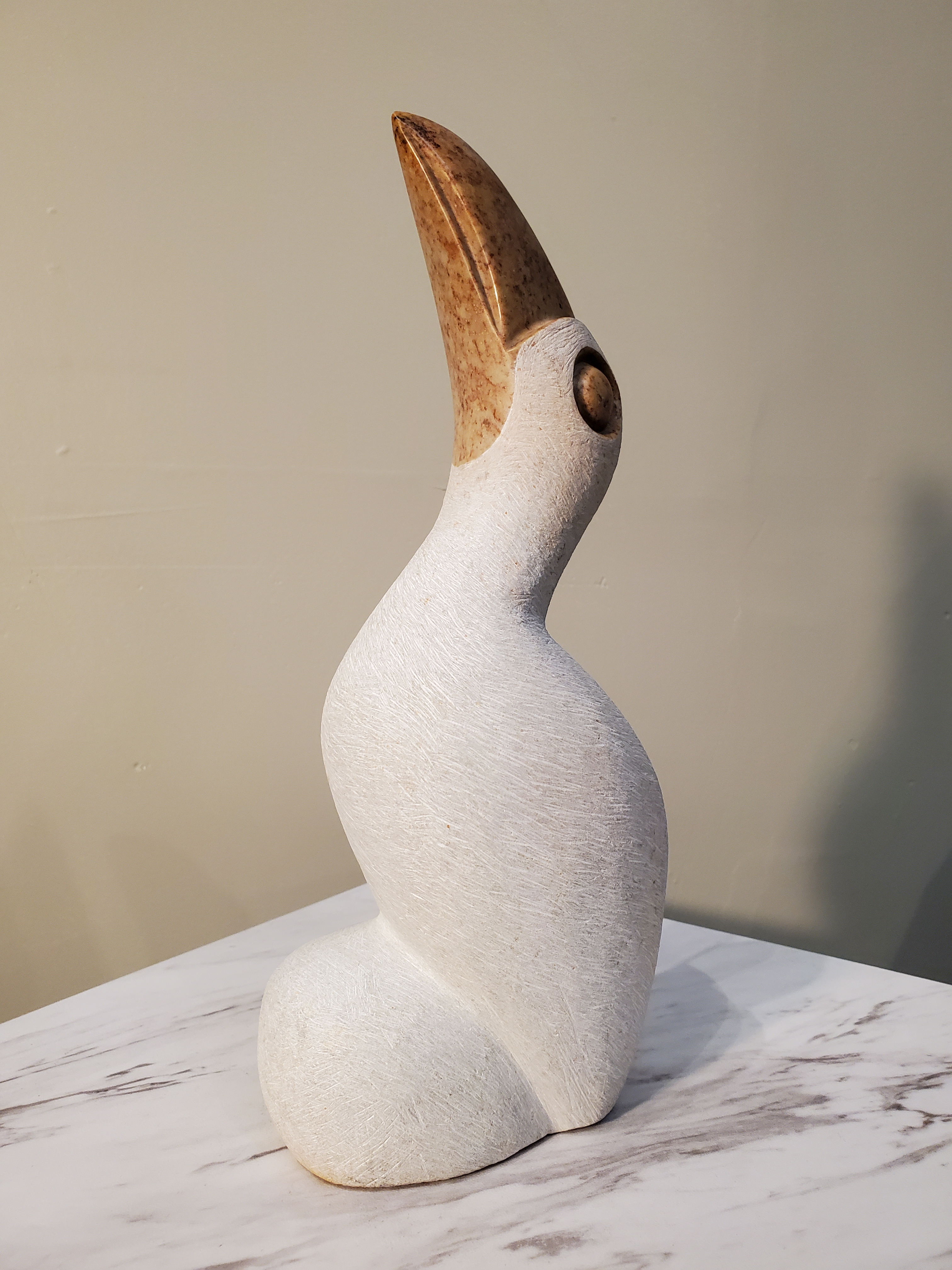 A beautiful perched white pelican looking inquisitively upward. This bird was sculpted from a single piece of opal stone. The white bird is accentuated by a bronze colored beak and eyes. Colors included: White, Bronze.